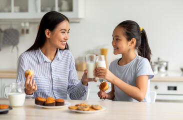 happy asian girl and her mother drinking milk while sitting in kitchen, clinking glasses and enjoyin