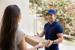© fizkes - Male courier in blue uniform and cap hands over sealed cardboard package to smiling woman at entrance of her residential home, engaged in positive and friendly interaction. Doorstep delivery services