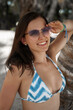 © aleonovs - A young, fit woman with long brown hair, light skin, and pink sunglasses smiles playfully under the tropical sun. She wears a blue bikini and leans against a tree, with a sunlit beach