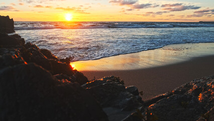 Poster - Beautiful natural landscape with sandy beach  with big stones during sunset.  Sun over horizon and Rolling ocean waves to the rocky shore while sunset.  Summer tourist destination in Portugal. Travel