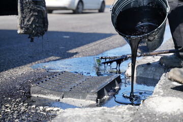 Fototapete - Close-up of hot bitumen being poured from metal bucket onto fresh asphalt around water drainage cover to create seal at highway road construction site