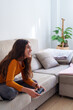 © Javier Zayaz - Smiling female gamer enjoying videogame, gripping controller while relaxing on cozy living room sofa