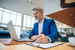 © zinkevych - Businessman in suit sitting in automobile dealership office using laptop