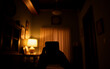 © YiuCheung - Dimly lit room interior, featuring a silhouette of a person holding a phone in the foreground. Background reveals furniture and curtain under soft light.