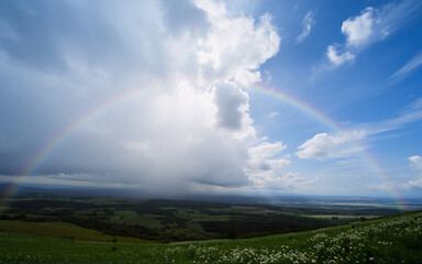 Poster - A colorful rainbow sweeps across a lush green landscape beneath a sky filled with fluffy white clouds and patches of bright blue.