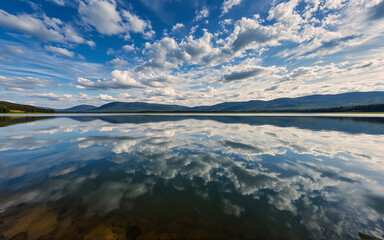 Poster - Serene landscape featuring a calm lake reflecting fluffy white clouds and a bright blue sky. Distant mountains are visible on the horizon under a sunny day.