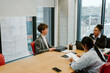 © Drobot Dean - A male intern and a female employee are sitting at a table and smiling while talking, with a female employee sitting next to them looking at papers, in the office, during an internship