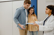 © Drobot Dean - A pregnant woman stands with her partner in a medical clinic hallway, smiling as a female doctor shows them medical documents. The couple listens attentively during their prenatal appointment.