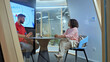 © wkproduction - In a modern office setting, a man and woman sit at a table, engaged in a discussion. The man wears a red shirt, while the woman is in a pink top. A large screen behind them displays a diagram.
