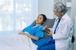 © ArLawKa - An elderly Asian female doctor is writing on a clipboard with a young female patient in a hospital room.