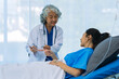 © ArLawKa - An elderly Asian female doctor is writing on a clipboard with a young female patient in a hospital room.