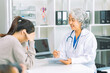 © ArLawKa - An elderly Asian female doctor is writing on a clipboard with a young female patient in a hospital room.