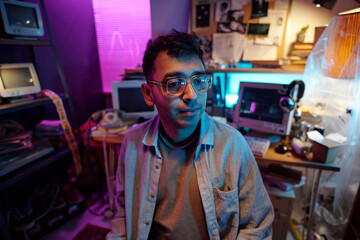 Medium closeup of young programmer wearing glasses sitting in dimly lit room with retro devices and notice board in background