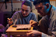 © TrueFrame Collective - Medium closeup of young woman soldering motherboard on wooden stool while her male friend assisting