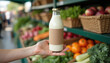 © KateBo - Milk bottle mockup. Blank labeled milk bottle in hand at local food market — packaging mockup surrounded by organic vegetables for natural dairy branding visuals.