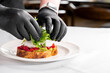 © pavel siamionov - A chef wearing black gloves prepares a gourmet toast with poached eggs, avocado spread, roasted red peppers, and fresh arugula on a white plate, showcasing vibrant colors and textures.