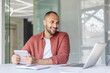 © Liubomir - A smiling man sits at a desk, using a laptop and holding a tablet. Natural light floods the modern workspace, enhancing the scene