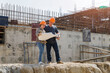 © skarie - Two construction workers discuss project details on-site, using a tablet and blueprints to plan the next steps amid the bustling construction environment.