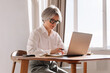 © Viktor Solomin/Stocksy - Senior woman working on laptop at dining table in modern office
