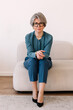 © Viktor Solomin/Stocksy - Mature woman with silver hair and glasses sitting on a modern sofa