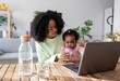 © Westend61 - Single working mother helping daughter drinking water from baby cup sitting with laptop at table