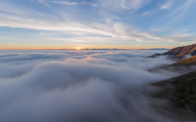 Aufkleber - A stunning aerial view captures a landscape where thick, white clouds blanket the valleys beneath mountain peaks during a colorful sunset with a gradient sky.