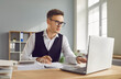 © Studio Romantic - Portrait of young attractive business man accountant working at the desk with a pile of financial documents on his workplace looking at the laptop monitor screen at the office and typing.