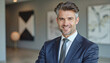 © f_bossa - A portrait headshot photo of a friendly professional CEO executive business worker: A handsome, graying man in a dark suit and tie smiles confidently at the camera in a modern, minimalist lobby.