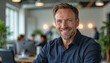 © f_bossa - A portrait headshot photo of a friendly professional CEO executive business worker: A smiling, middle-aged man with graying hair sits in a modern office, looking directly at the camera with a war