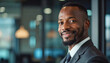 © f_bossa - A portrait headshot photo of a friendly professional CEO executive business worker: A confident Black businessman in a suit smiles warmly at the viewer, with a blurred, modern office setting in t