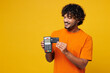 © ViDi Studio - Young happy Indian man he wears orange t-shirt casual clothes hold wireless modern bank payment terminal to process acquire credit card isolated on plain yellow background studio. Lifestyle concept.