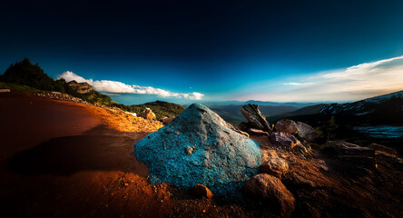Poster - Vibrant mountain landscape features a textured rock in the foreground under a clear blue sky with distant trees and peaks illuminated by warm light.