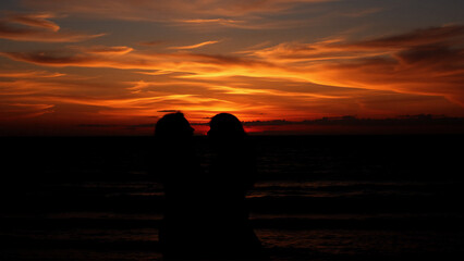 Poster - Fiery sunset paints the horizon with vibrant orange and red hues as dark silhouettes stand against the ocean's edge.  The sky glows with warmth.