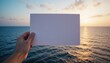 © Chirawan - Hand Holding Blank White Paper Against Ocean Sunset Background