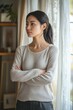 © Elena - Asian woman with her hands on her hips, looking out a window with curtains drawn. She is wearing a cozy sweater and has long hair tied up.