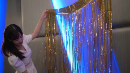 Poster - Asian Woman in White Dress Decorating Wall with Gold Streamers Under Blue Light, Creating Festive and Elegant Atmosphere