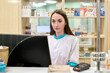 © TRAVELARIUM - Young female pharmacist with long brown hair, wearing white coat, focuses on computer screen. Bright, organized pharmacy background with shelves of medicines