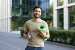 © Tetiana - Portrait of a smiling young Indian male athlete standing outside, holding a mat and a bottle of water, looking at the camera