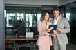 © insta_photos - Two busy older professional corporate business executives man and woman wearing suits working on digital project plan holding tablet technology device having discussion standing in office at meeting.