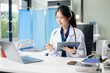 © Nuttapong punna - Confident young female doctor in white medical uniform sit at desk working on computer.