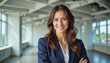 © f_bossa - Modern portrait headshot of a friendly ceo executive business worker: A confident businesswoman with wavy brown hair smiles directly at the camera while standing in a modern, open-plan office space.
