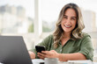 © Vergani Fotografia - Cheerful Latin businesswoman in her 30s wearing green button-up shirt using smartphone while working on laptop in bright modern office with urban skyline view through large windows