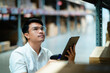 © KANGWANS - A man in a white shirt is looking at a tablet while standing in a warehouse. He is holding a barcode scanner in his hand