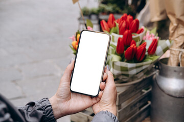mockup woman's hand holding using smart phone with white blank screen pov at flower street shop, mob