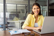 © Stock 4 You - Portrait at camera smiling latin female marketing manager, professional it specialist working at laptop computer sitting at desk in modern office. Cheerful young woman employee using pc for business
