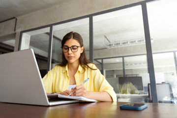 Focused young female woman student watching webinar video online for studying. Latin hispanic girl in casual clothes, eyeglasses learning business lessons writing notes sitting in office. Copy space