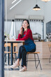 © PRIME STOCK LAB - Cheerful woman wearing red sweater and blue apron sits at wooden table in modern cafe, smiling warmly.