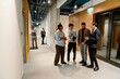 © Drobot Dean - A young, overweight Indian woman and a young White man, who are in their 20s, laughing together with their older male colleagues in an office hallway, as other people work in the background