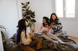 © Drobot Dean - A diverse group of four young women in their late 20s, laughing and having fun while sitting on a bed together in a minimalistic white bedroom, doing makeup and chatting