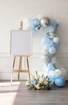 A white, blank canvas on an easel, a blue and silver balloon arch, a white wall background